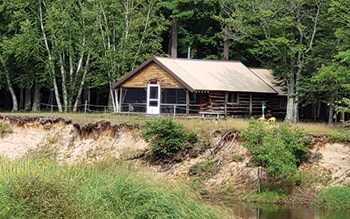 Brown log cabin on the river on summer day with green grass and plants covering the bank that the cabin overlooks.