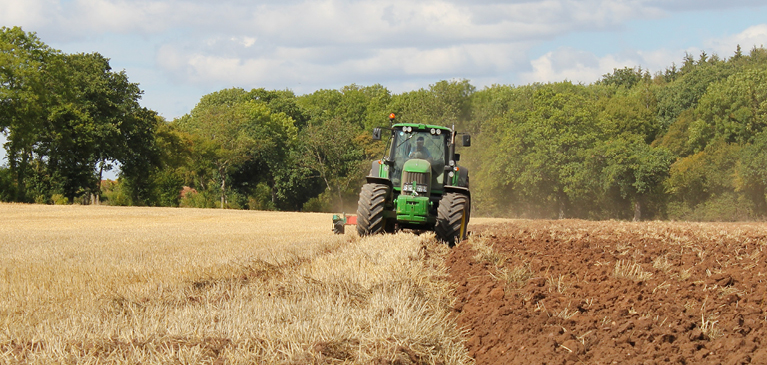 Green medium-sized tractor plowing fields in the summer with blue sky and white clouds.