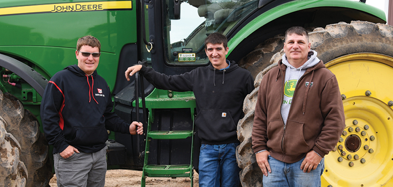 Two young farmers standing beside their uncle in front of a large John Deere tractor