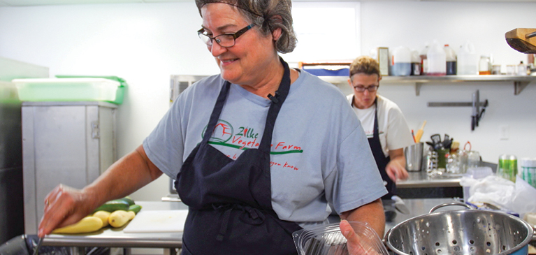 Woman with apron and hairnet preparing food in commercial kitchen with another woman in the background helping