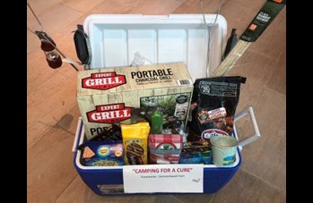 Camping for a Cure basket with cooler holding s'mores sticks, grill, lantern, food, and camping gear.