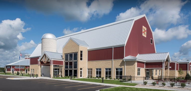 Outside photo of the Farm Wisconcin Discovery Center, a large red and brick barn with many buildings with a yellow barn quilt block in corner of barn on a sunny summer day.