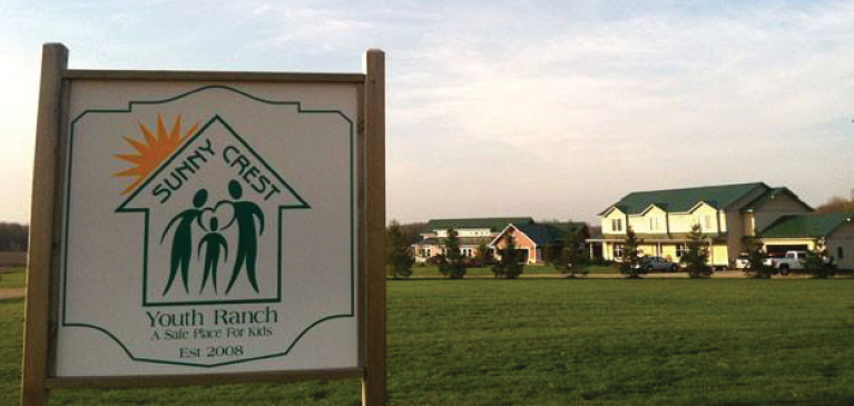 Sign at Sunny Crest Youth Ranch on sunny day with two of the housing units in view