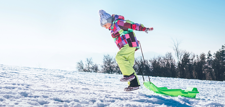 Young girl in colorful coat and green snowpants leaping while pulling plastic sled up snowy hillbank.