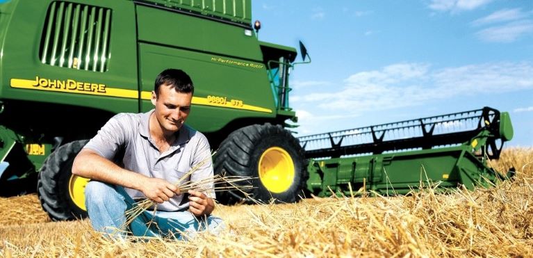 Young male farmer kneeling in front of John Deere combine in a hay wheat field during harvesting on sunny day