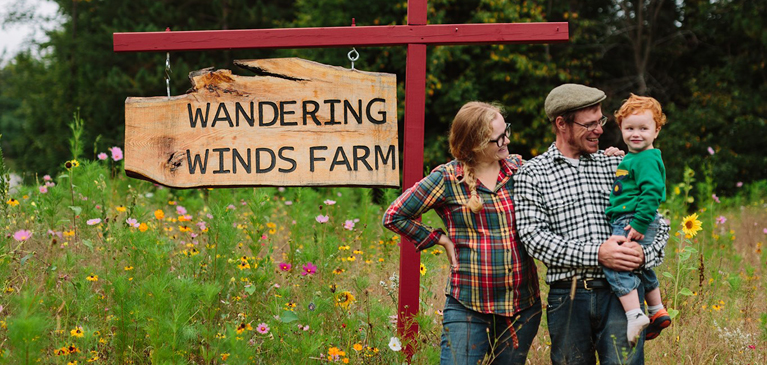 Woman in red flannel shirt standing next to her husband in green flannel holding their son by their custom farm sign in a meadow.